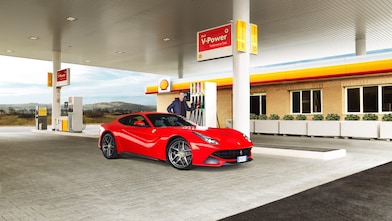 A red Ferrari sitting on a Shell station forecourt with a man leaning on a petrol pump